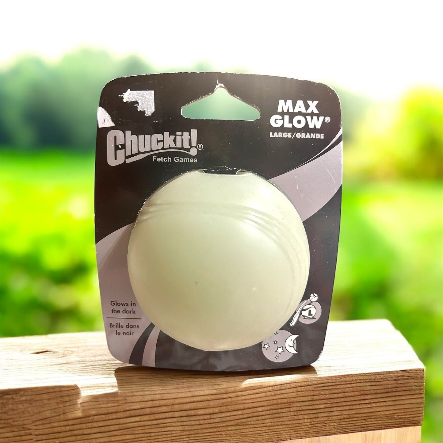 Chuckit! Max Glow dog ball packaging on a wooden surface with a blurred green outdoor background